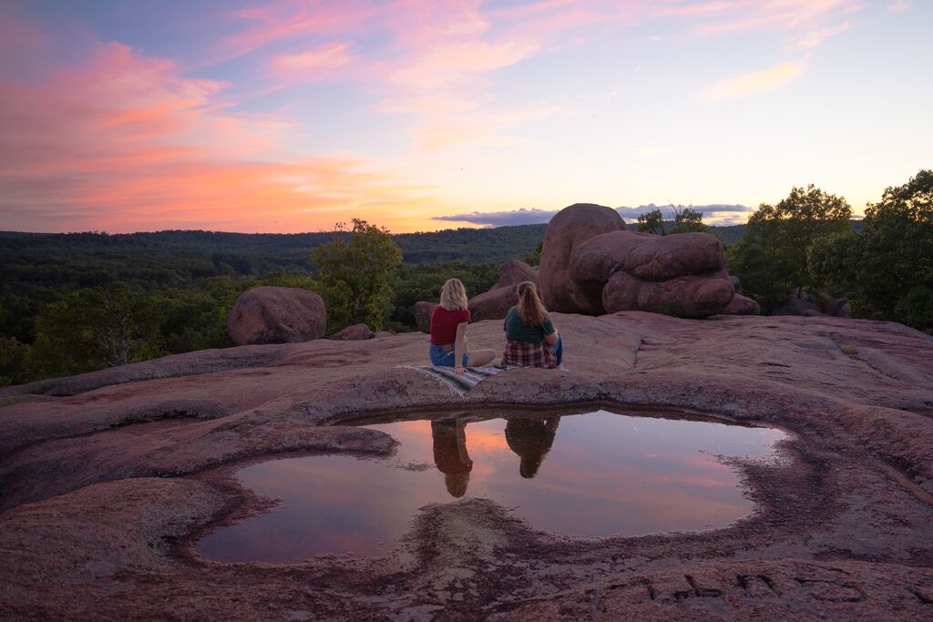 Two people sit on top of a large rock formation during sunset with their reflection visible in a pool of water behind them.