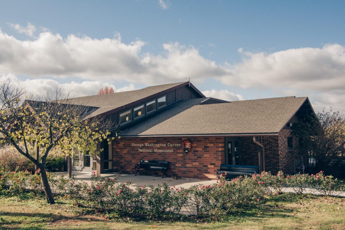 A large brick building with greenery, black benches, National Park Service Arrowhead logo, and a description: George Washington Carver National Monument.
