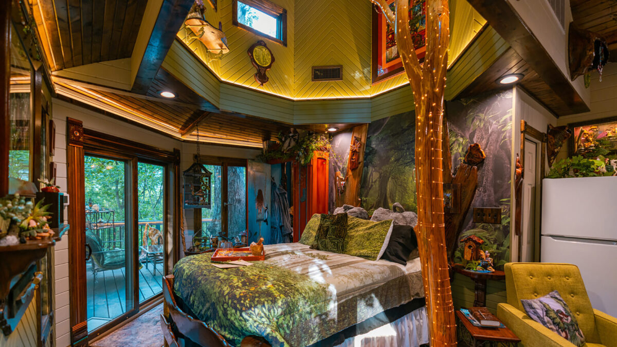 An interior view of the Enchanted Forest Treehouse. There is a large bed, vaulted ceilings, a tree covered in fairy lights, and a sliding glass door revealing a deck and lush forest. The room is decorated whimsically, as though fairies live there.