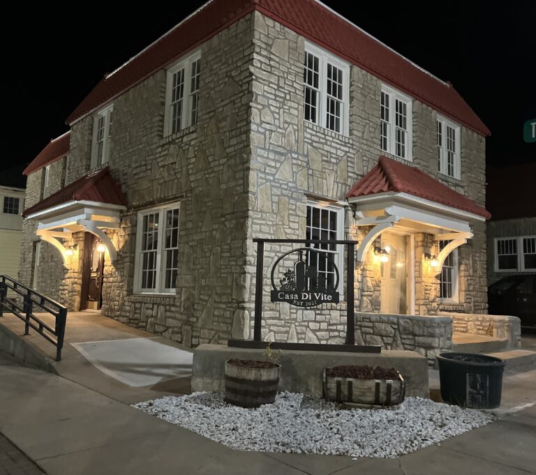 The exterior of a grey brick building with a red roof and lit entryways.