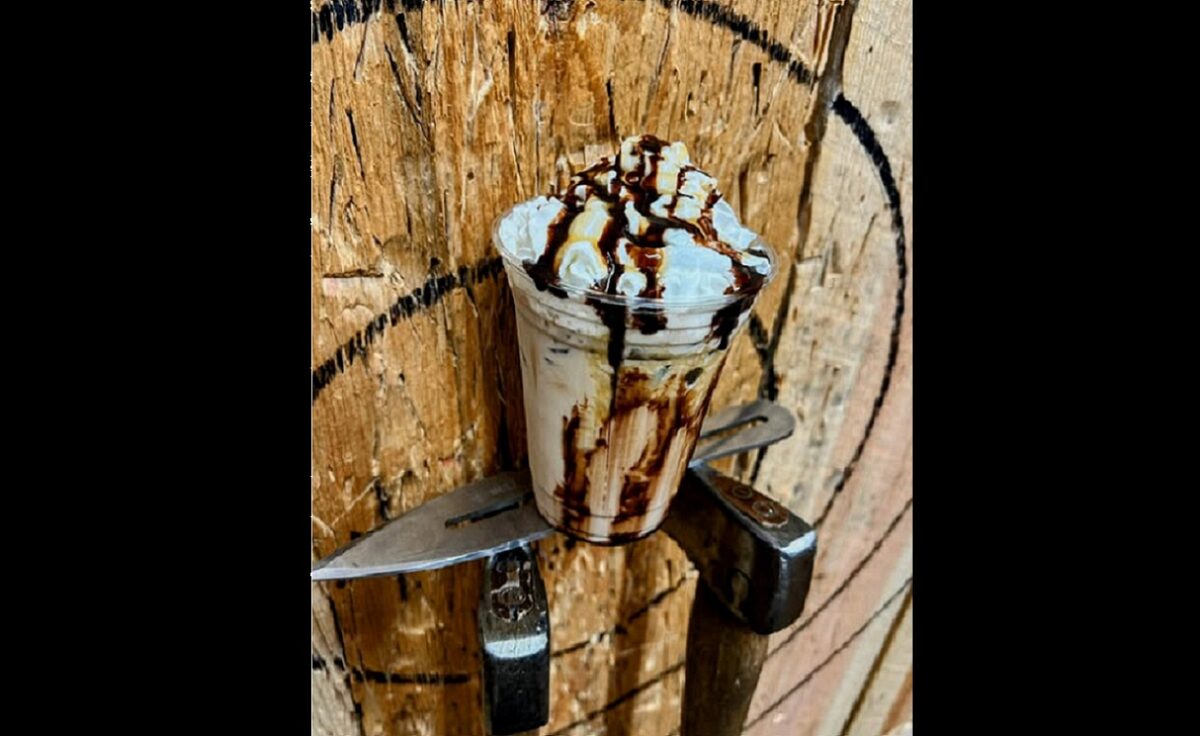 A milk shake atop the head of an axe sunk into the wood of a target.