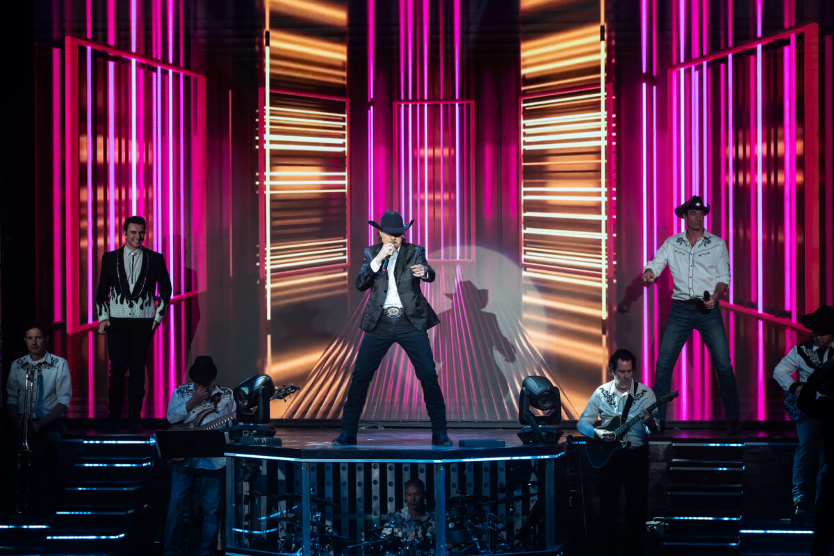 Three performers on stage before a colorful background.