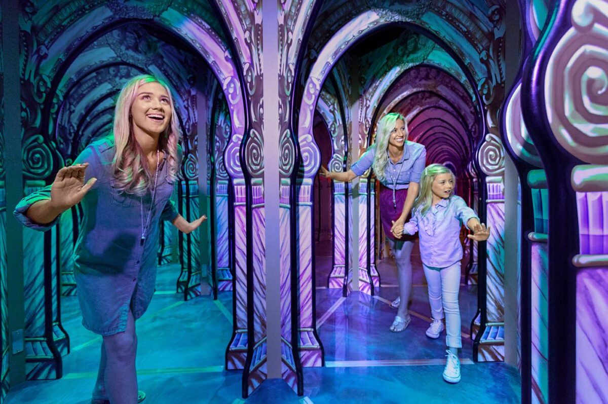 A family explores a vibrant, mirror-filled maze with colorful, glowing arches. A young girl leads the way, holding hands with her smiling mother, while another woman follows close behind, all appearing excited and engaged in the experience.