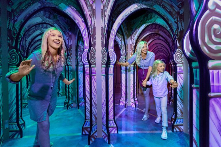 A family explores a vibrant, mirror-filled maze with colorful, glowing arches. A young girl leads the way, holding hands with her smiling mother, while another woman follows close behind, all appearing excited and engaged in the experience.