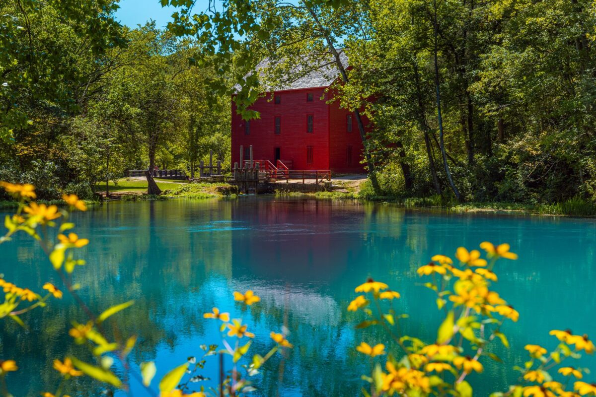 A rich red mill sits nestled in lush green trees with a vibrant turquoise spring in the front. There are bright yellow wildflowers in the foreground.