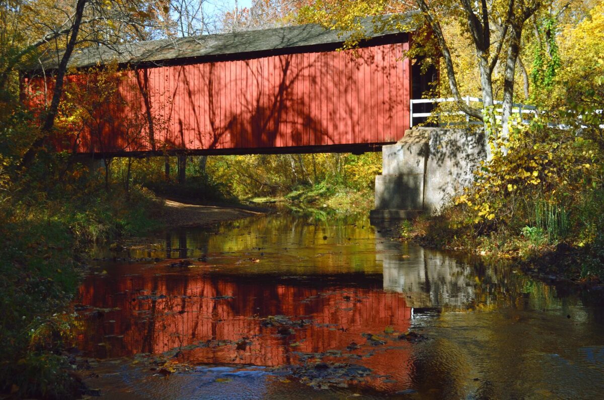 A red covered bridge is perched above a shallow creek. It's vibrant reflection can be seen in the trickling water.