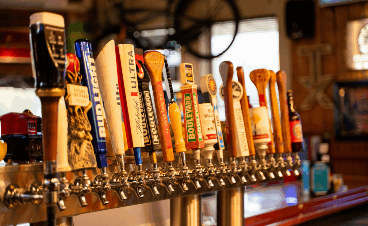 A long row of polished beer taps at Frisco’s bar, featuring a variety of brands including Boulevard, Michelob Ultra, Bud Light, Angry Orchard, and more, with a rustic backdrop of vintage décor and neon lights.