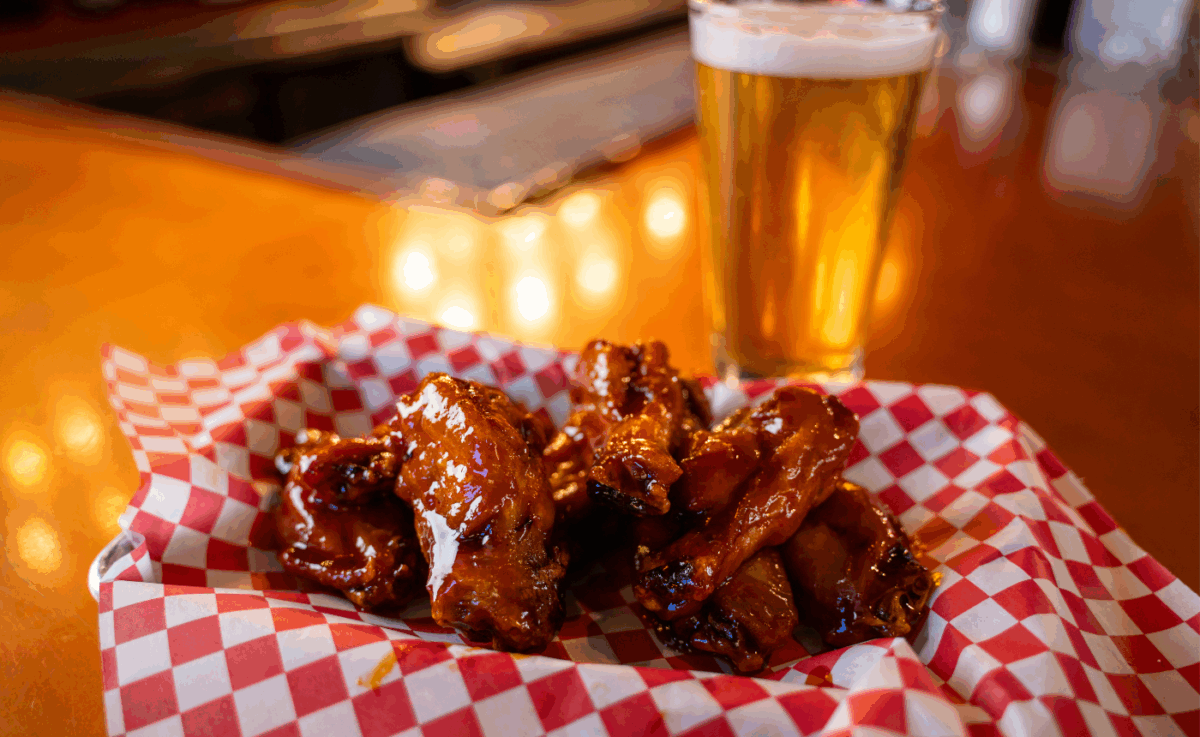 A red-and-white checkered basket of saucy glazed chicken wings served fresh at Frisco’s bar, paired with a golden pint of beer on a polished wood counter with warm lighting in the background.