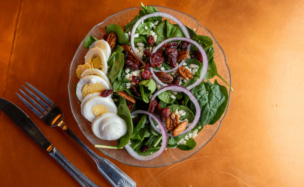 A fresh spinach salad served on a clear glass plate, topped with hard-boiled egg slices, red onion rings, chopped pecans, dried cranberries, and crumbled cheese, accompanied by a fork and knife on a copper-toned table.