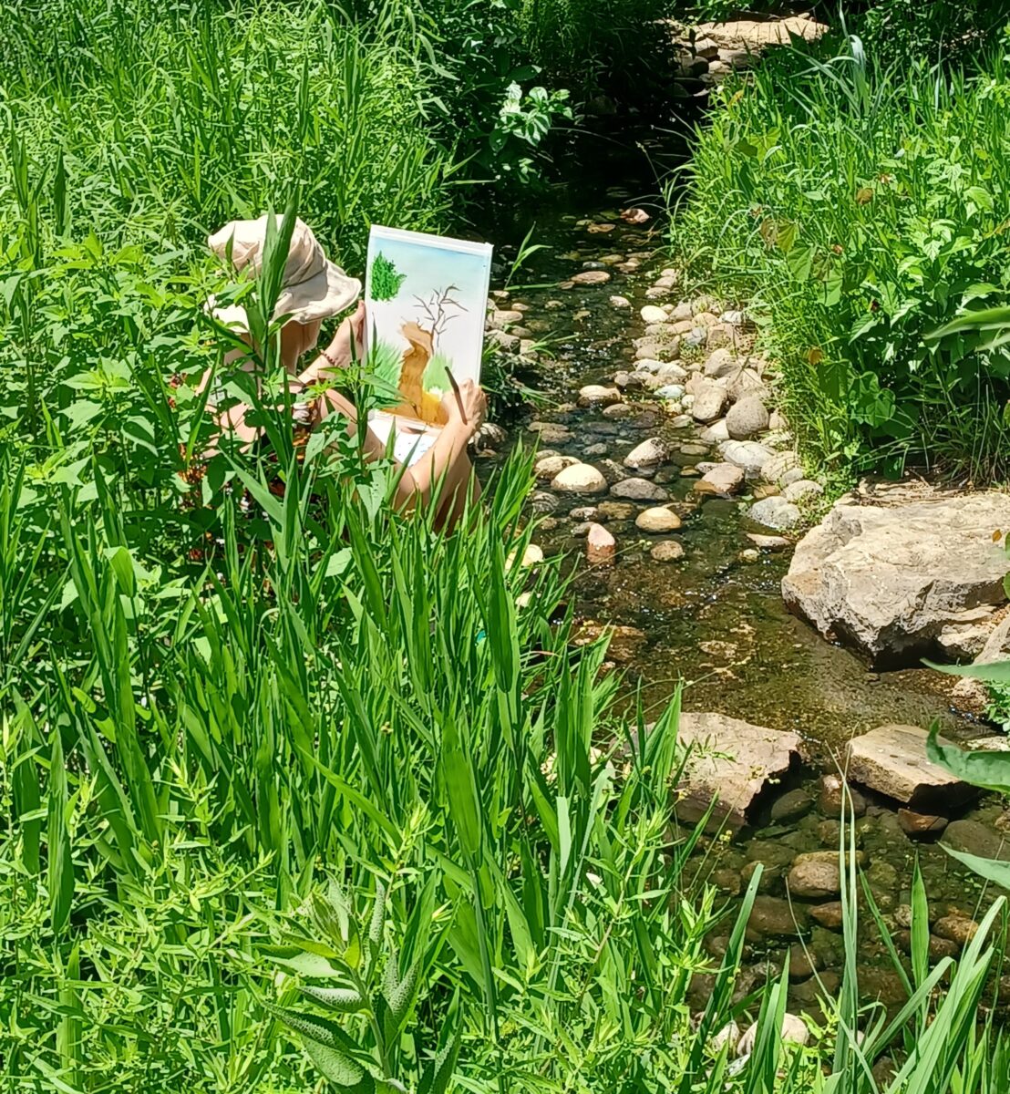 Person sits next to small stream painting flowers.