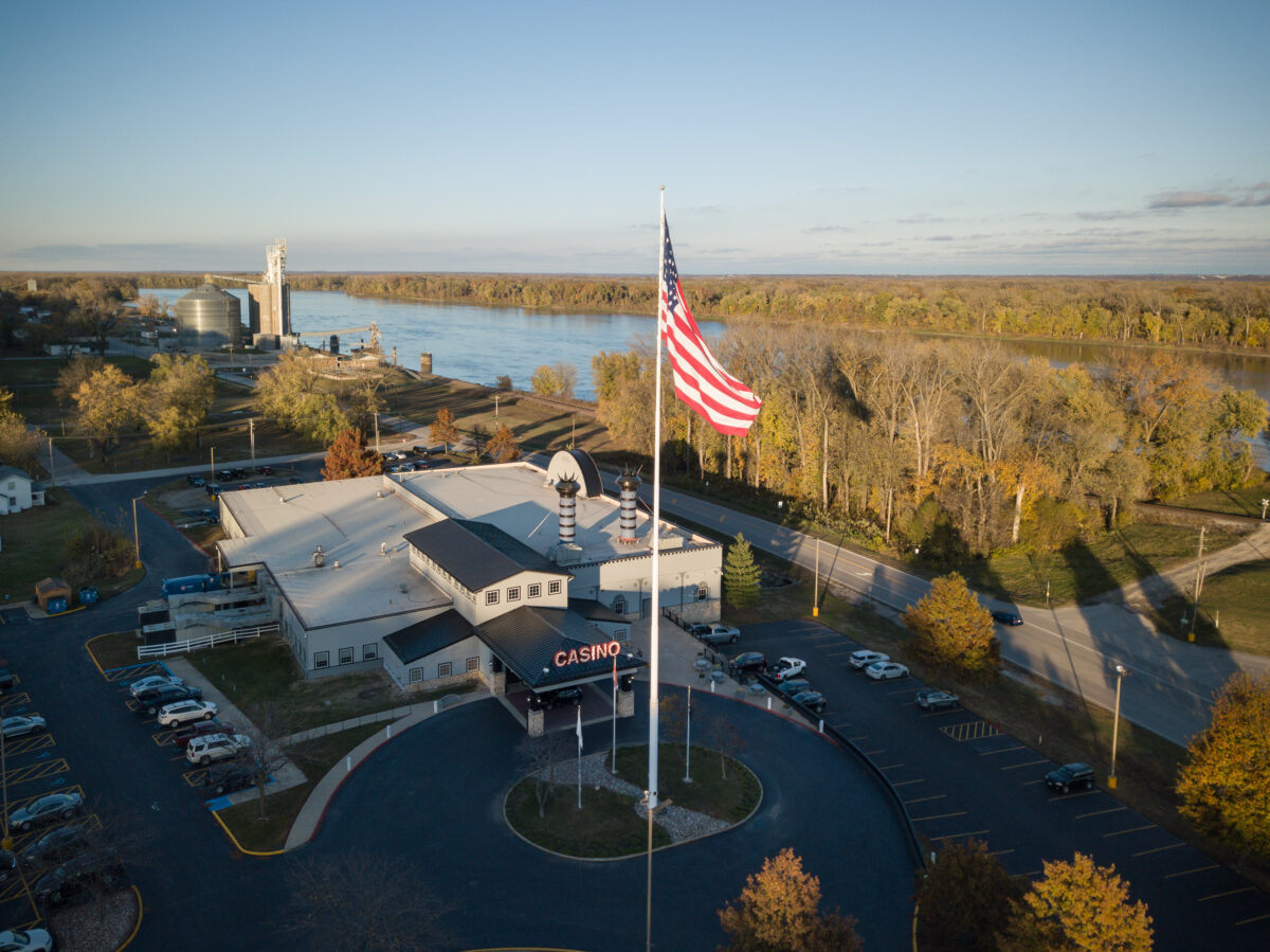 An American flag at full staff raised over a courtyard near a body of water and trees.