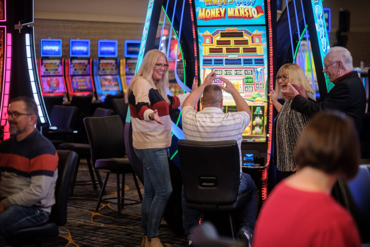 A group of people smile, laugh and cheer as a man sits and plays at a slot machine.