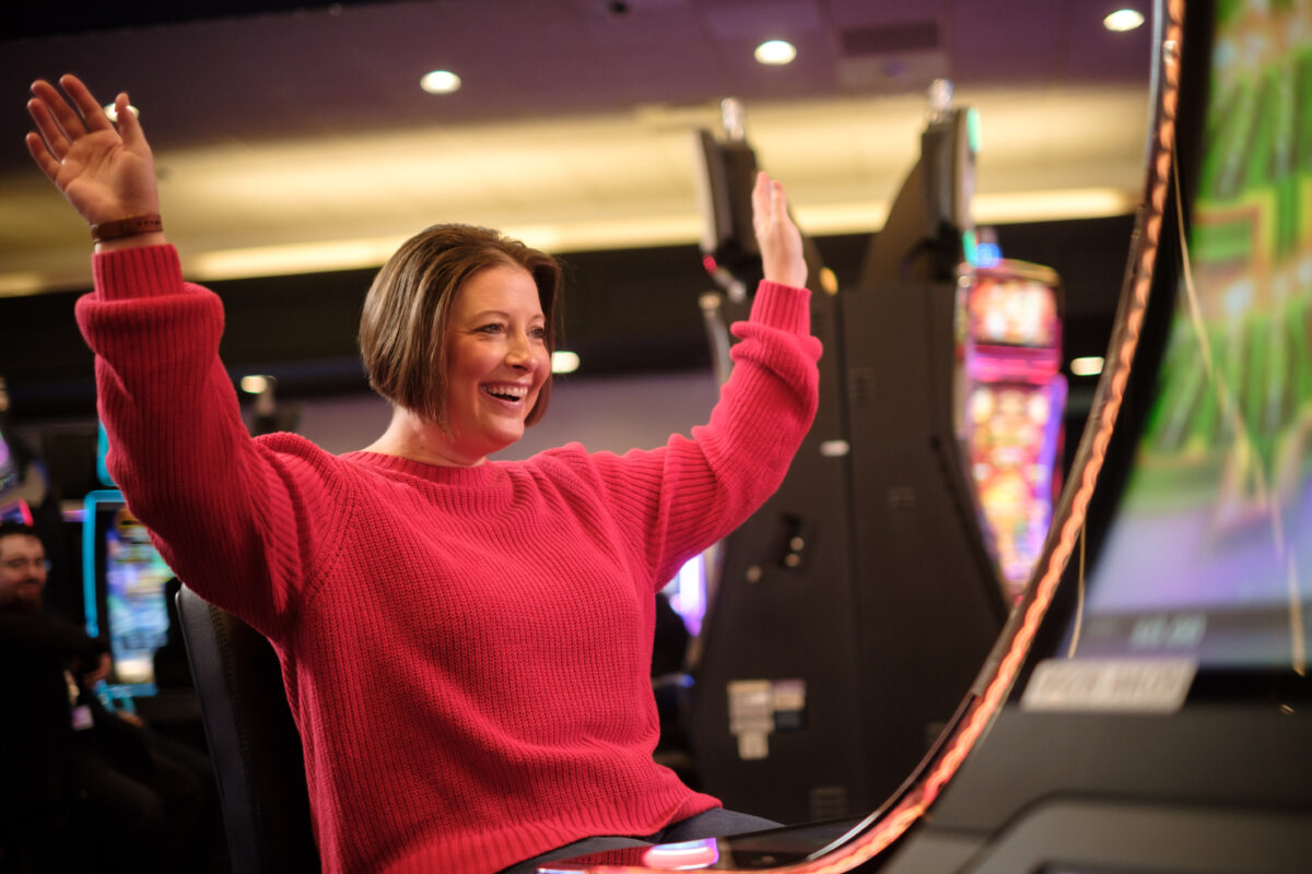 A woman in a red shirt cheers with her hands up and smiles while playing a slot machine.