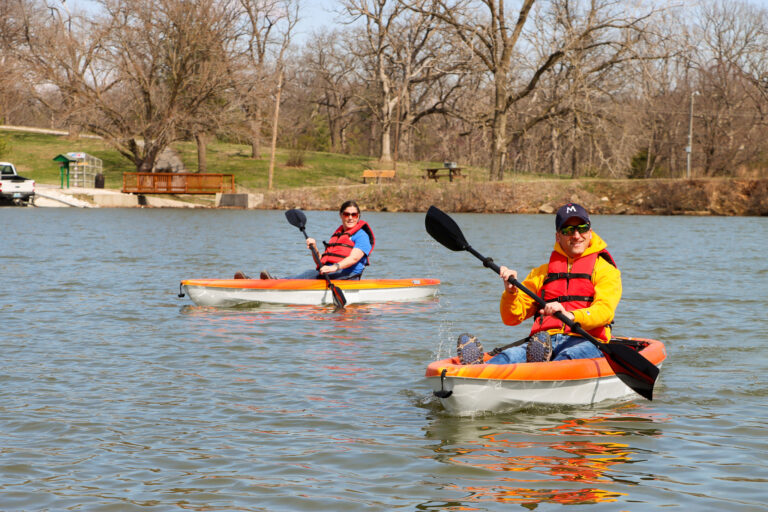 Two people kayak on a lake wearing orange life vests.
