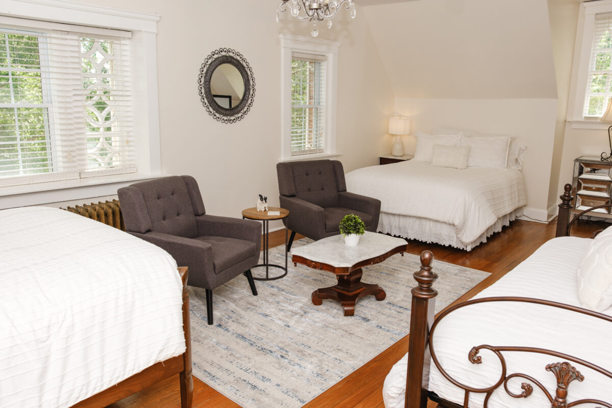 Bright bedroom with multiple beds, white bedding, two chairs, chandelier, and large windows.
