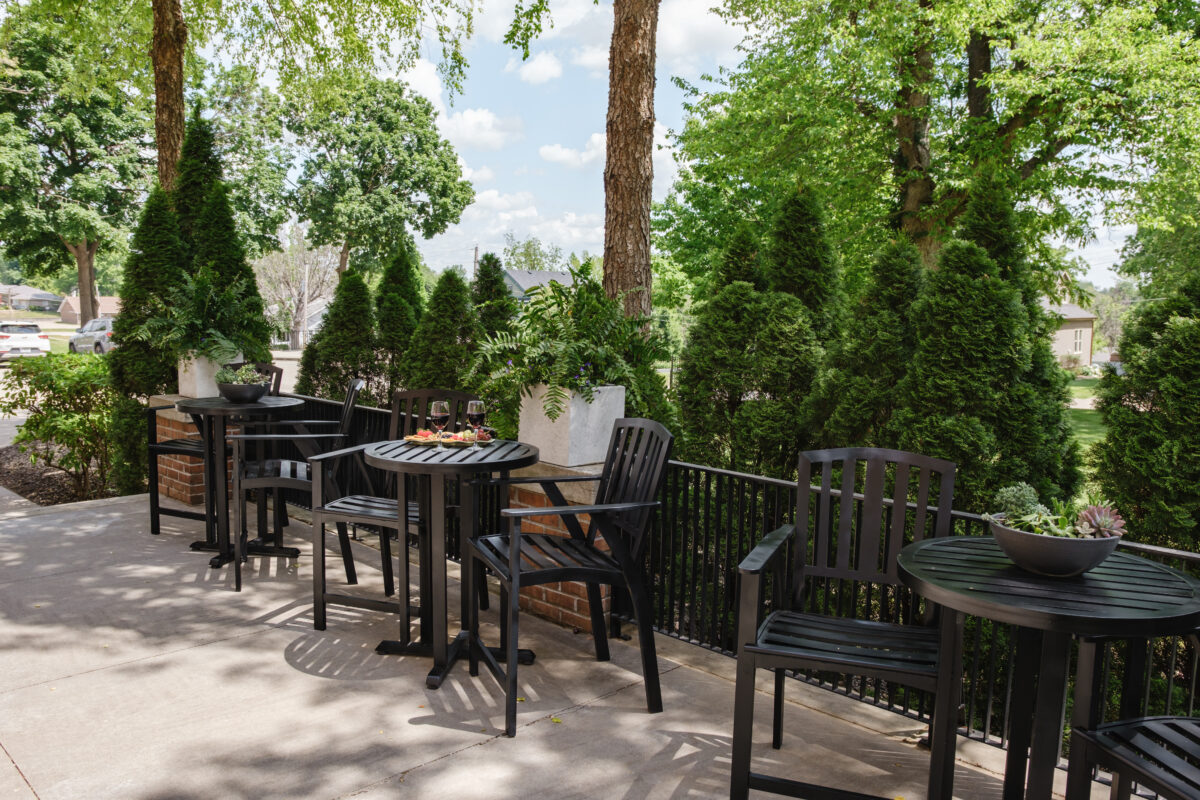 Outdoor patio with black tables and chairs surrounded by greenery and trees.