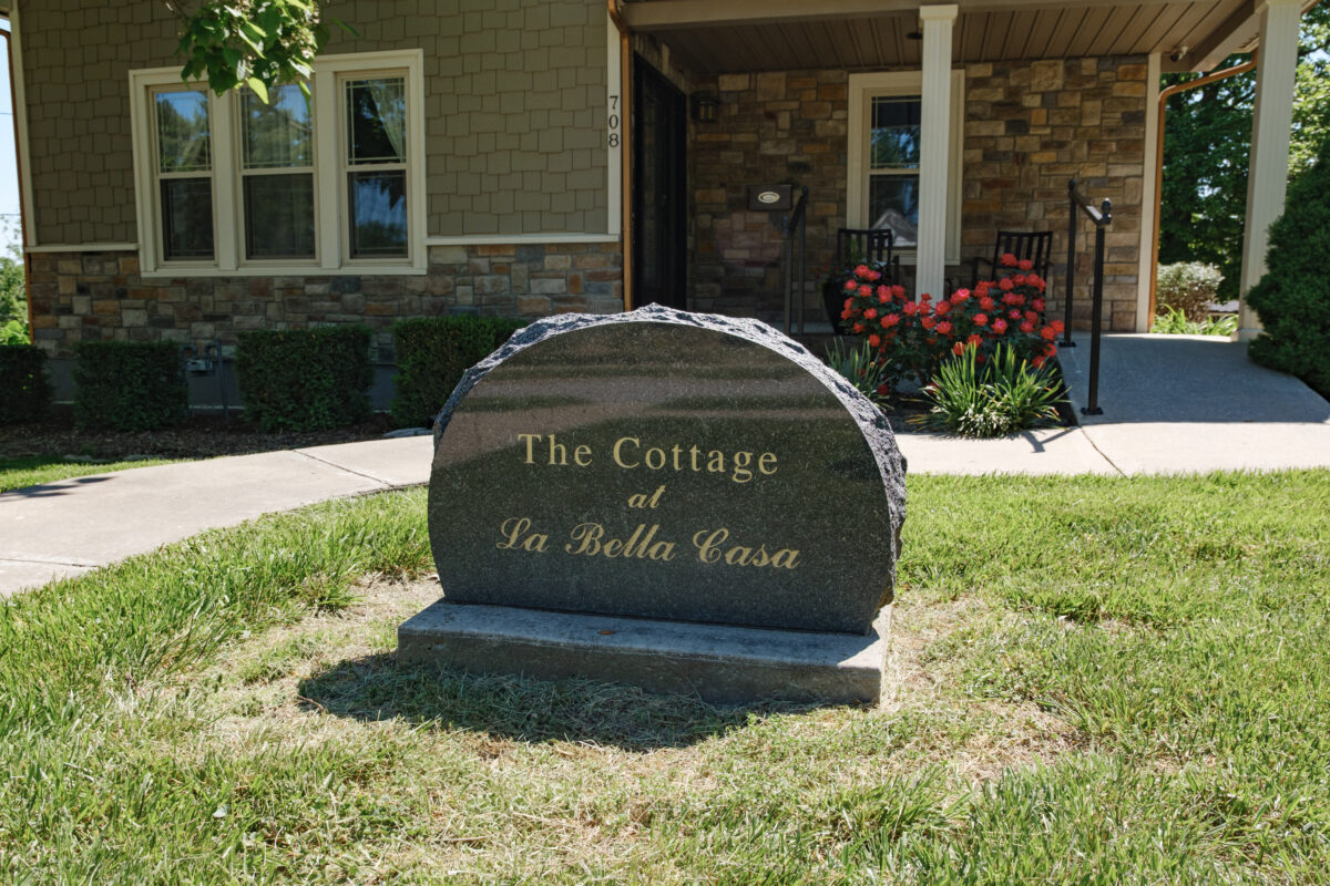 Stone sign reading “The Cottage at La Bella Casa” in front of a landscaped entrance with flowers and covered porch.