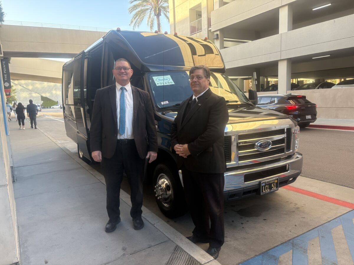 Two people in suits pose outside of a large black vehicle.