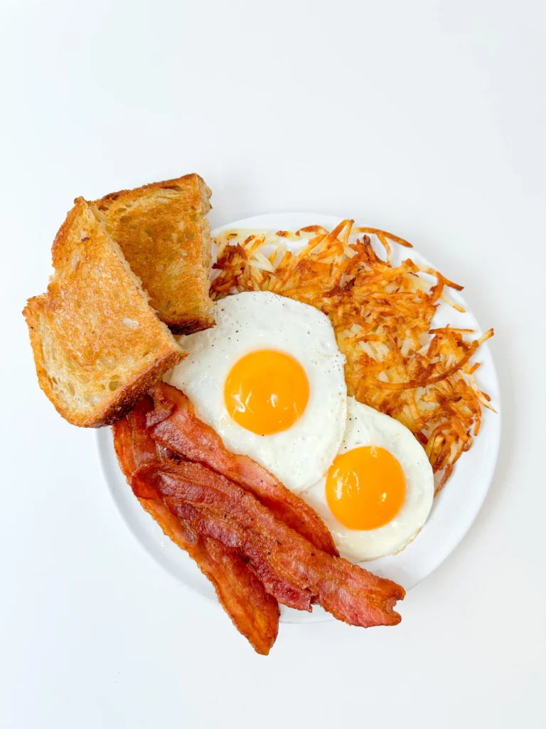 A plate of bacon, sunny-side-up eggs, hashbrowns and toast against a white background.