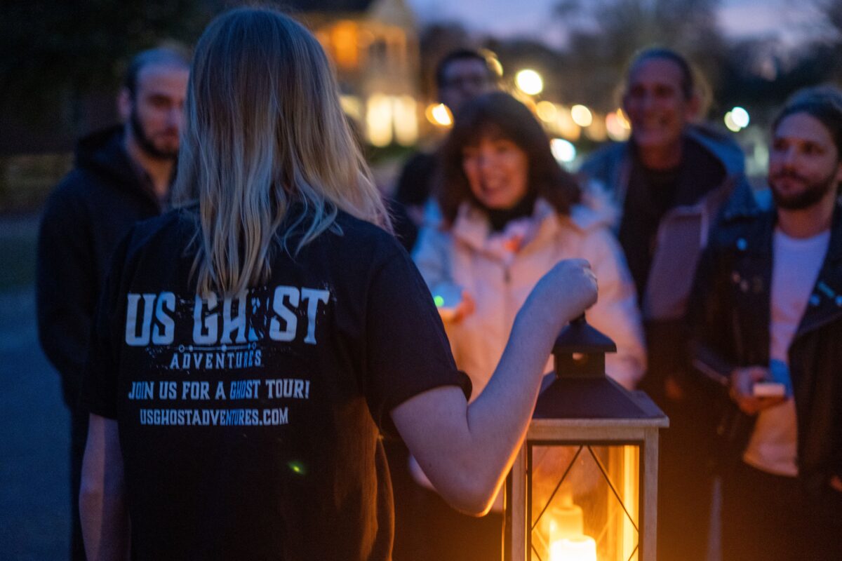 A ghost tour employee with her back to the camera and a lantern in her hand addresses attendees of the ghost tour.