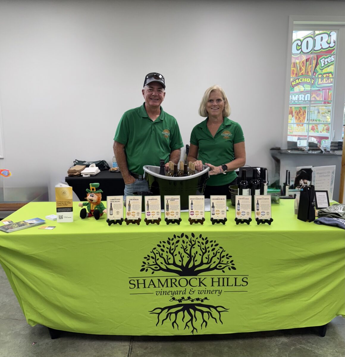 Two people pose behind a table with a green cloth that reads "Shamrock Hills Vineyard and Winery." The table holds cards detailing their various wines.