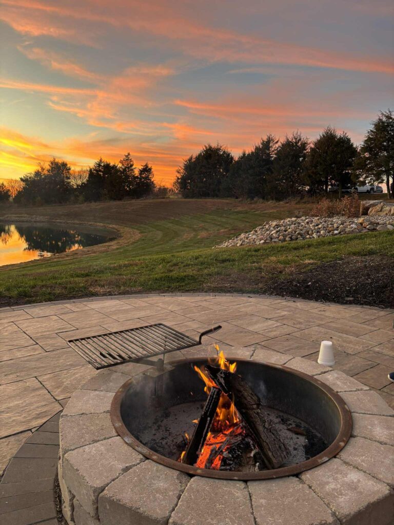 A lit outdoor fireplace on a circular pathway surrounded by trees, rocks and a pond at sunset.