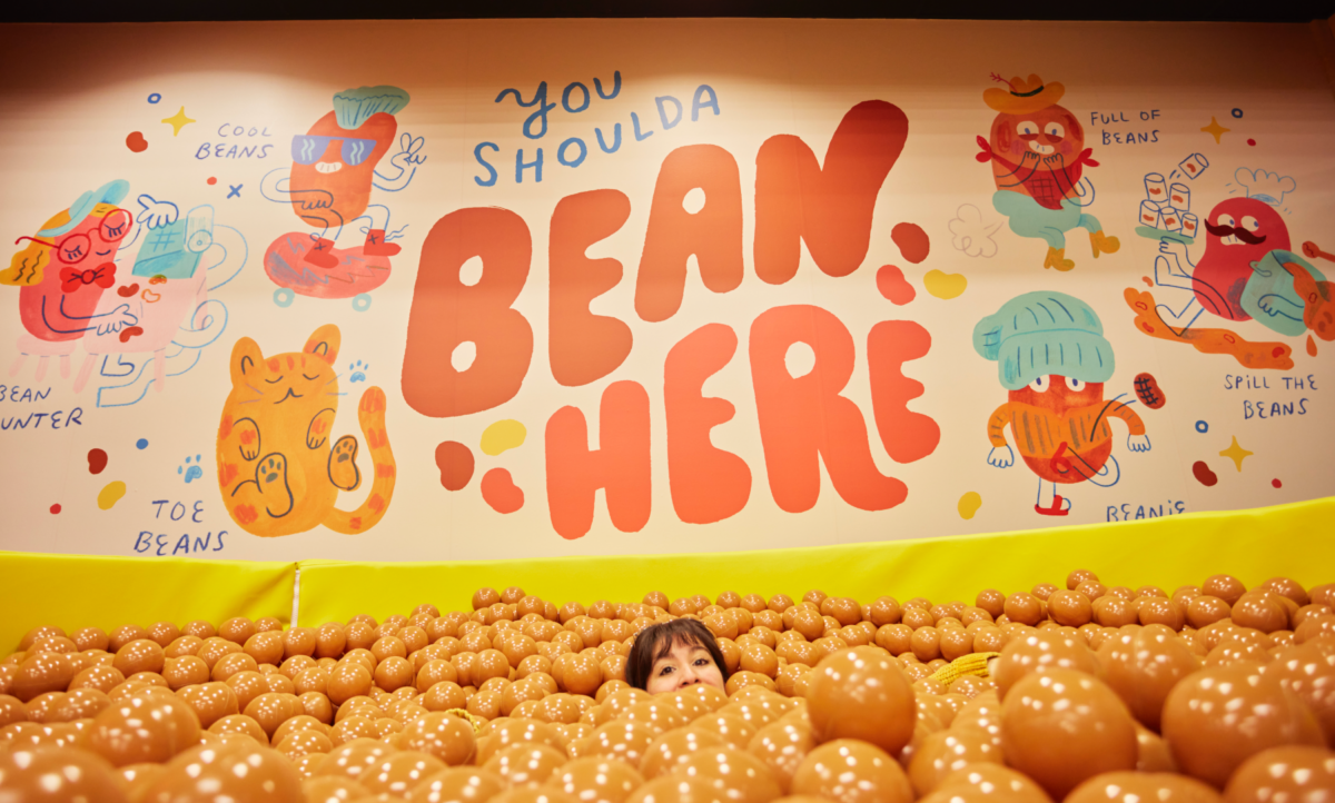 A person’s head pokes out of a bean pit filled with brown ball pit balls below a mural that says You Shoulda Bean Here.
