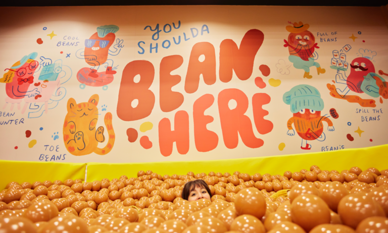 A person’s head pokes out of a bean pit filled with brown ball pit balls below a mural that says You Shoulda Bean Here.
