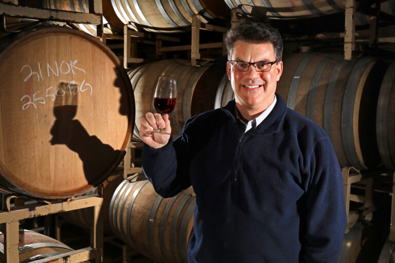 A man in a shirt and sweater raises a glass of wine to the camera.