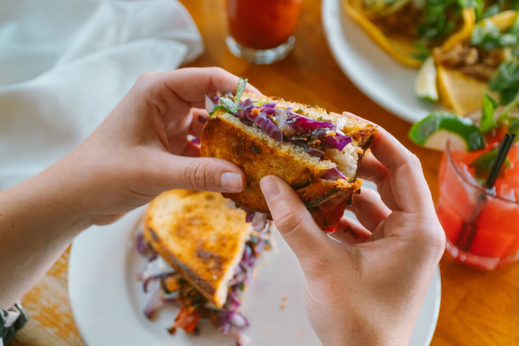 Hands hold a sandwich on toasted bread with a fun cocktail in the background.