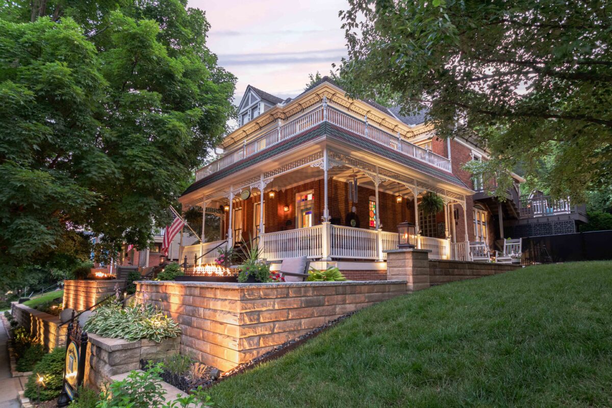Historic 1885 brick home with a wraparound porch, Victorian trim, and warm evening lighting at the Main Street Inn in Parkville, Missouri.