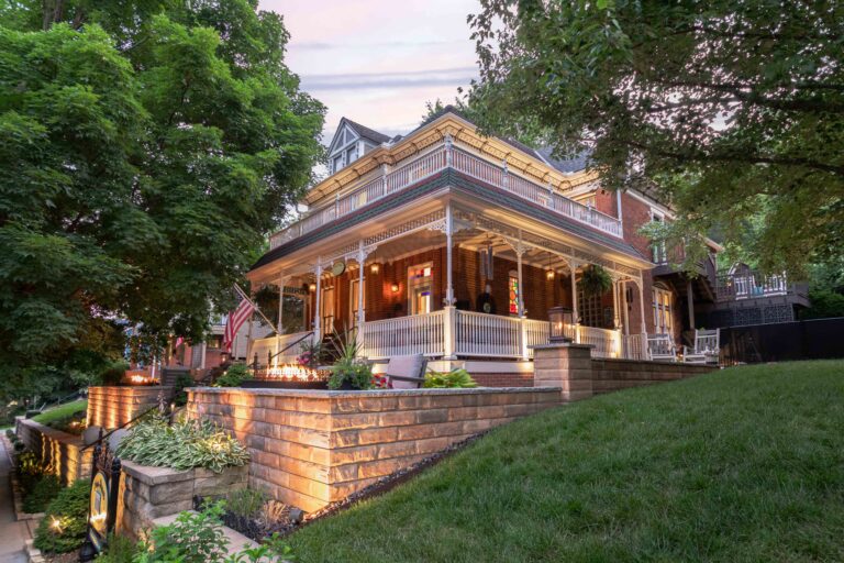 Historic 1885 brick home with a wraparound porch, Victorian trim, and warm evening lighting at the Main Street Inn in Parkville, Missouri.
