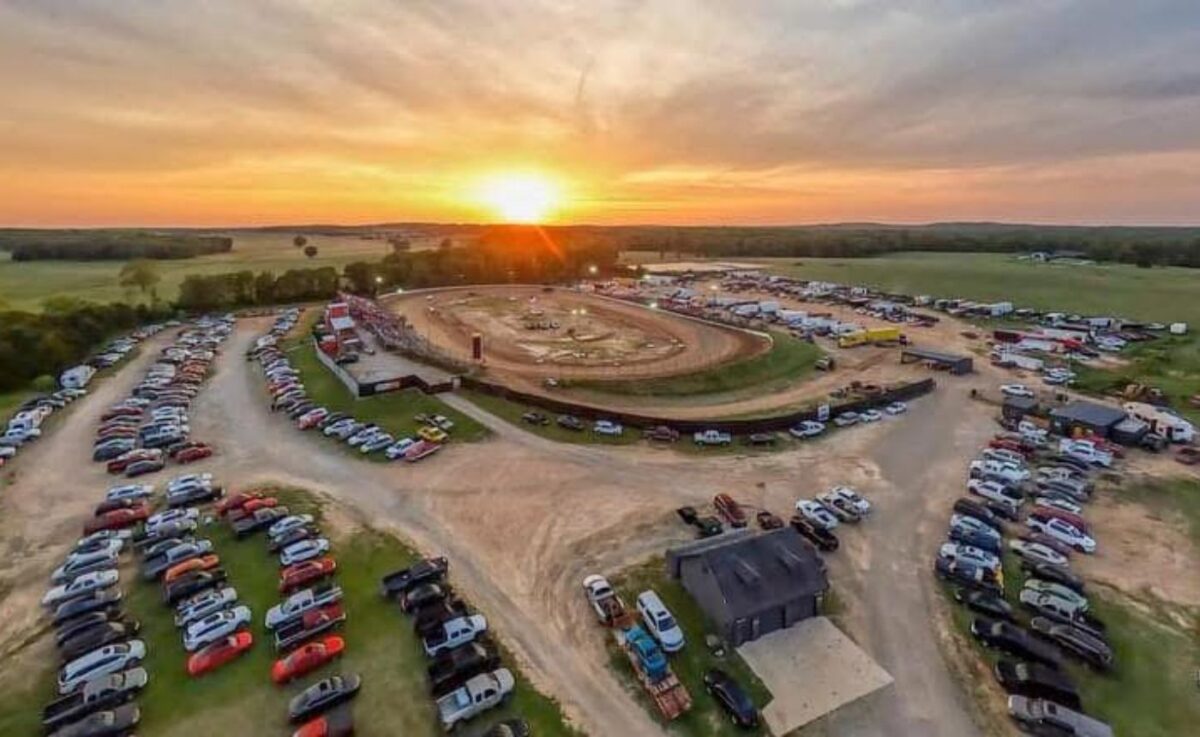 An aerial view of Midway Speedway in Lebanon, Missouri, capturing a dirt oval racetrack at sunset. The track is surrounded by green fields, with dozens of cars and trucks parked in organized rows around the perimeter and near the main entrance building. The setting sun on the horizon creates a bright glow and casts long shadows across the facility.