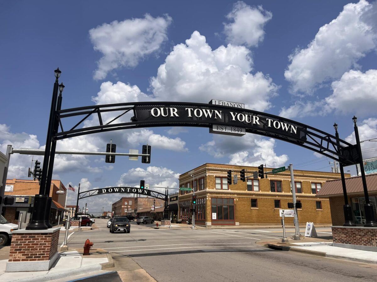 An eye-level shot shows the downtown area of Lebanon, Missouri, on a bright, sunny day. A large, black metal arch spans across a wide, gray asphalt street, with the words "LEBANON," "OUR TOWN YOUR TOWN," and "EST. 1849" written on it. Behind it is another smaller, similar arch with the word "DOWNTOWN" on it. On the right side of the street is a two-story, tan brick building with several windows and a green sign that reads "COMMERCIAL ST" and "JEFFERSON." A white car is parked on the left side of the street, and a black SUV is driving toward the camera. The sky is a vibrant blue, filled with fluffy, white clouds.