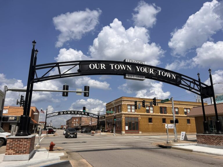 An eye-level shot shows the downtown area of Lebanon, Missouri, on a bright, sunny day. A large, black metal arch spans across a wide, gray asphalt street, with the words "LEBANON," "OUR TOWN YOUR TOWN," and "EST. 1849" written on it. Behind it is another smaller, similar arch with the word "DOWNTOWN" on it. On the right side of the street is a two-story, tan brick building with several windows and a green sign that reads "COMMERCIAL ST" and "JEFFERSON." A white car is parked on the left side of the street, and a black SUV is driving toward the camera. The sky is a vibrant blue, filled with fluffy, white clouds.