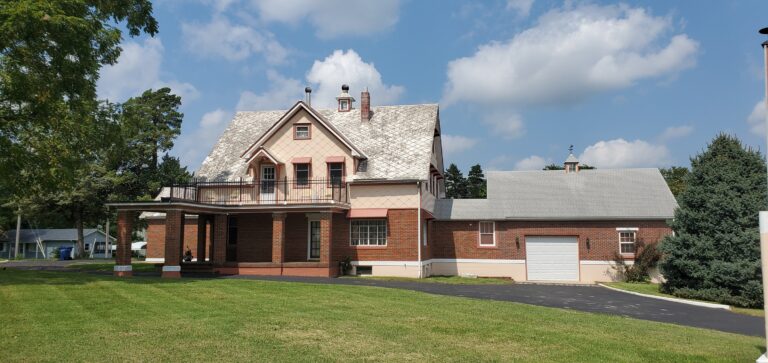 An outdoor, eye-level view of the Manor Inn on Route 66 in Lebanon, Missouri. This historic 1903 Victorian-style mansion features a tan and pink facade with a red brick ground floor. A large front porch and a second-story balcony with black railings face a paved driveway and a well-manicured lawn.