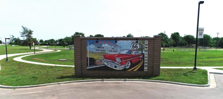An outdoor, eye-level view of a large Route 66 tile mural on a brick wall at Boswell Park in Lebanon, Missouri. It features a classic red 1957 Chevy cruising down the highway, flanked by a Missouri Route 66 shield and the slogan "Drive Our Town, In Your Town". A paved driveway leads to the monument, set against a backdrop of a winding walking path and a green park field
