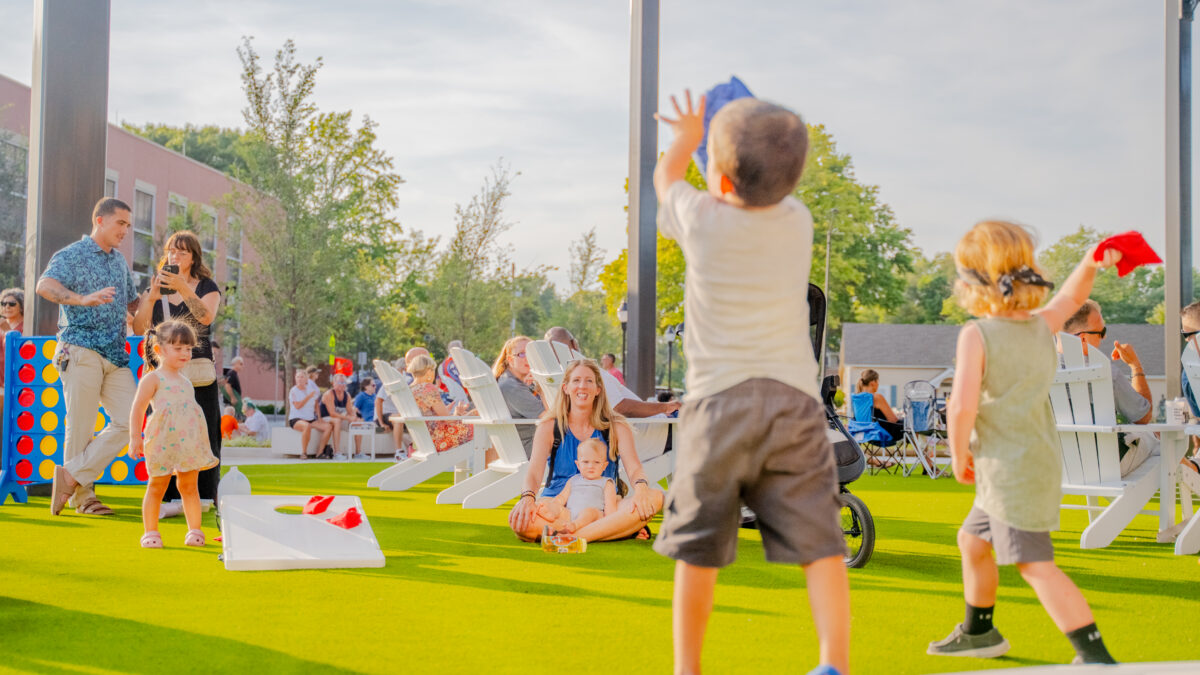 Children play cornhole outdoors during a farmers market event.
