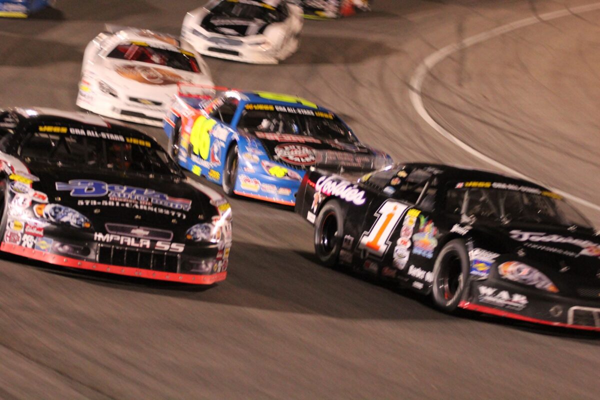 An action-packed shot of several late-model stock cars racing closely on an asphalt oval track at night. In the foreground, a black car numbered 87 with "Impala SS" on the front bumper and a black car numbered 1 with "Rackley WAR" branding battle for position. Close behind them is a blue and yellow car numbered 46, followed by two white cars in the background. The blurred background and the cars' leaning angles emphasize the high speed and intensity of short-track racing under stadium lights.