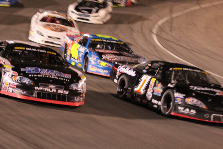 An action-packed shot of several late-model stock cars racing closely on an asphalt oval track at night. In the foreground, a black car numbered 87 with "Impala SS" on the front bumper and a black car numbered 1 with "Rackley WAR" branding battle for position. Close behind them is a blue and yellow car numbered 46, followed by two white cars in the background. The blurred background and the cars' leaning angles emphasize the high speed and intensity of short-track racing under stadium lights.