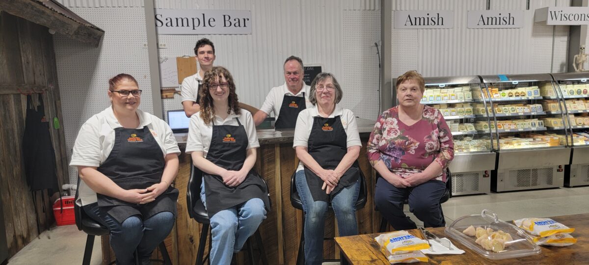 Several people in aprons pose in a cheese store.