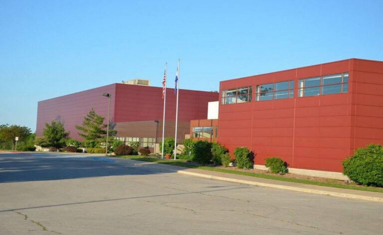 Exterior view of the Kenneth E. Cowan Civic Center in Lebanon, Missouri, showing its modern, maroon-colored rectangular buildings under a clear blue sky. Two flagpoles with the American and Missouri flags stand in front of the facility, which is surrounded by a paved parking area and manicured green shrubbery.