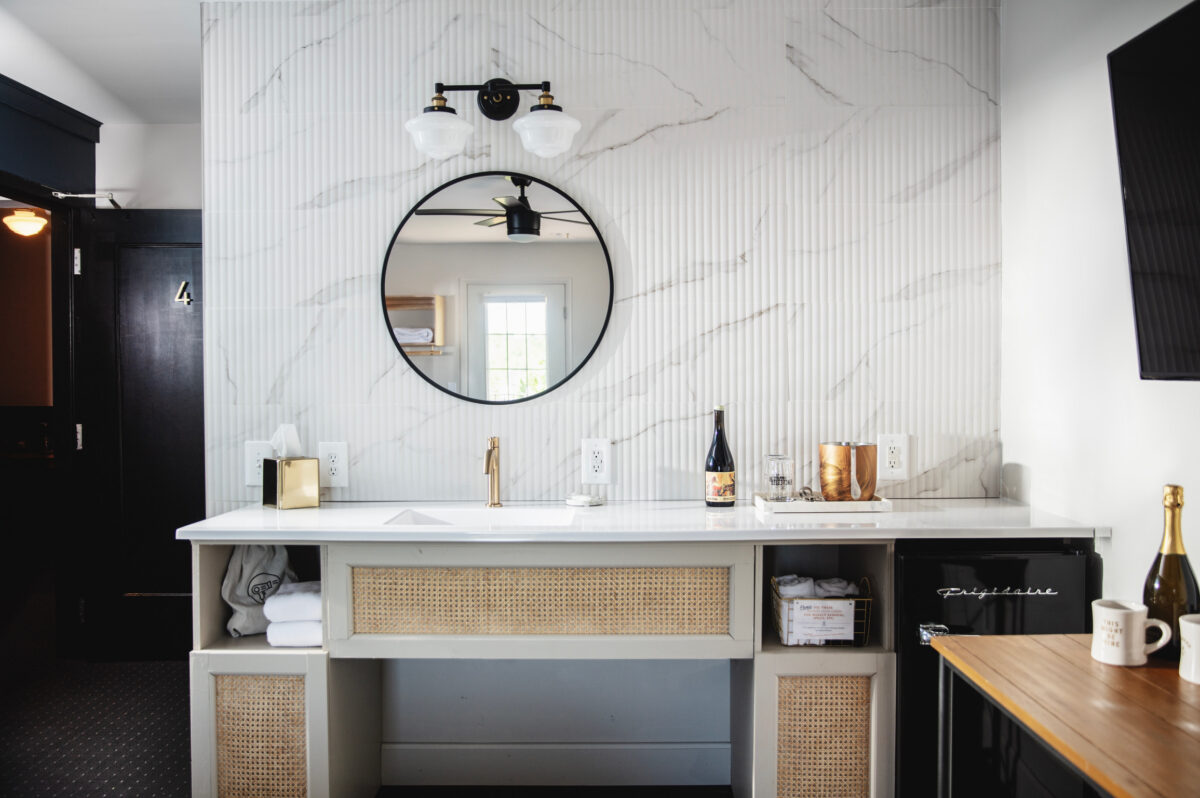 Modern bathroom vanity with natural cane accents, marble tile wall, a round mirror, and sleek white countertop. A warm ambiance with golden tap, wine bottle, and soft lighting.