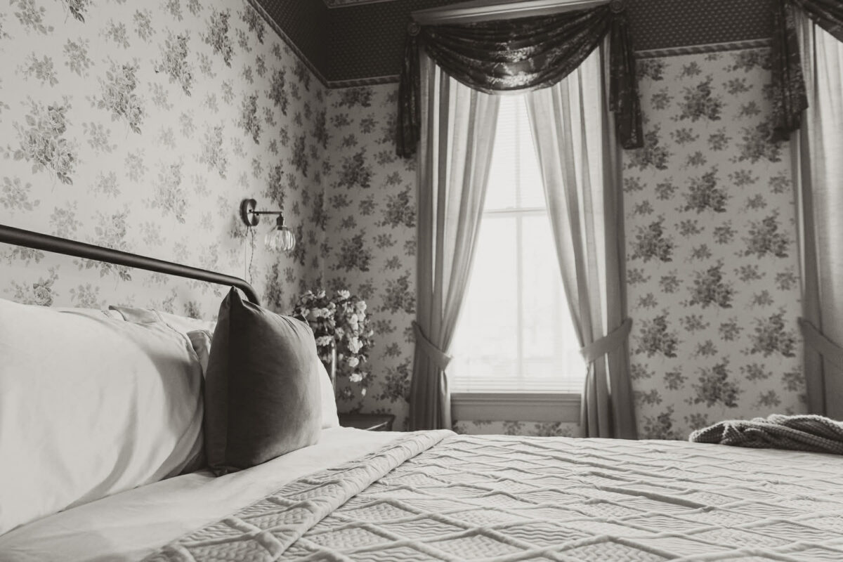 A black and white photo of a made bed and the wall and window on the other side of it.