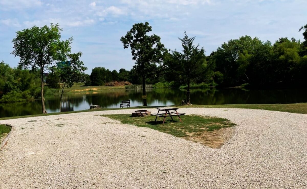 Gravel lakeside campsite featuring a wooden picnic table, fire pit, and view of a calm pond surrounded by lush green trees