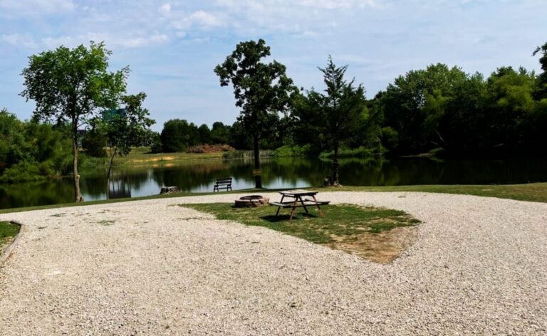 Gravel lakeside campsite featuring a wooden picnic table, fire pit, and view of a calm pond surrounded by lush green trees