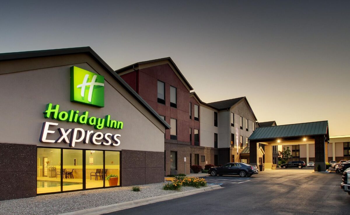 Exterior of a Holiday Inn Express hotel at dusk, featuring its illuminated logo on a modern, multi-story tan and brown building with a paved parking area.