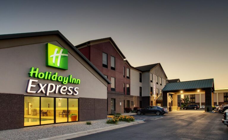 Exterior of a Holiday Inn Express hotel at dusk, featuring its illuminated logo on a modern, multi-story tan and brown building with a paved parking area.