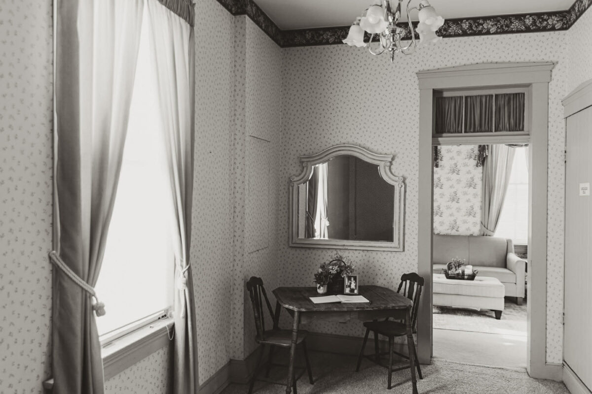 A black and white photo of a room with a window, mirror, table and chairs, and an open door to another room.