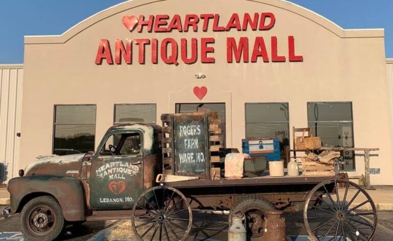 The exterior of Heartland Antique Mall in Lebanon, Missouri, featuring a large tan building with red signage. In the foreground, a vintage, weathered flatbed truck is parked, loaded with rustic antiques including a wooden crate labeled "Rogers Farm Ware, MO," old milk cans, and a blue trunk. The truck's door is hand-painted with the mall's logo and location.